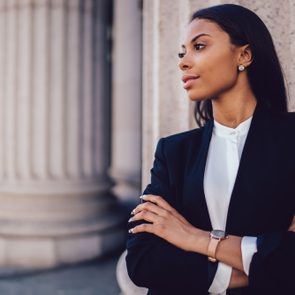 Female African American banker dressed in elegant black suit folding hands and looking on side standing against office building. Confident female entrepreneur with crossed arms looking on copy space