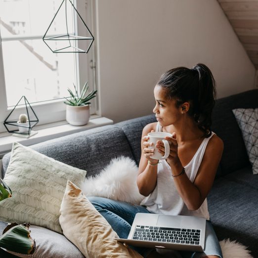 Young woman with laptop and cup of coffee sitting on the couch at home looking out of window