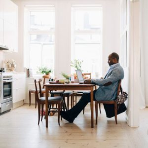 Full Length Of Mature Man Working On Laptop While Sitting In Kitchen At Home