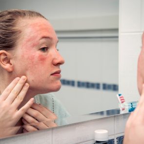 woman looking at skin reaction on face in bathroom mirror